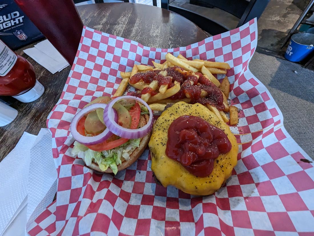 An open burger in a paper lined basket with fries, pickles, tomato and onion on the side.
