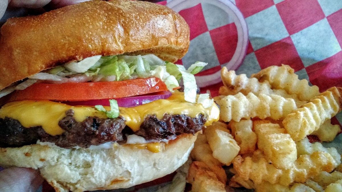 A cheeseburger with lettuce, tomato and red onion beside a pile of crinkle cut fries on checked paper.