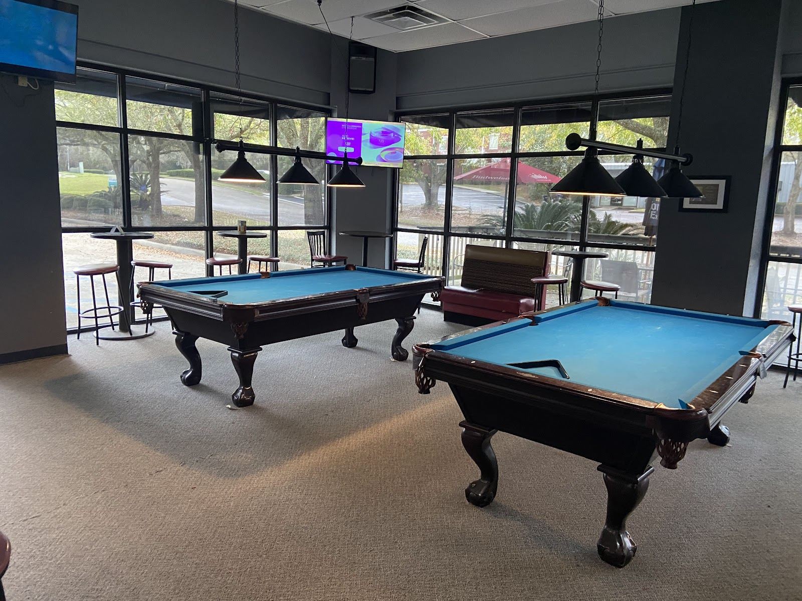 Two pool tables with bright blue felt under hanging lamps, beside tall windows looking onto the parking lot.