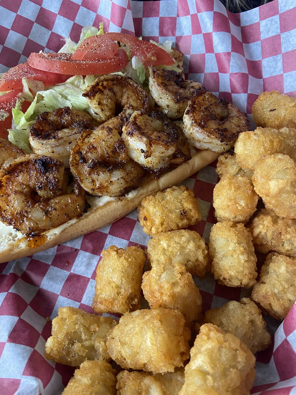 A long roll filled with seasoned shrimp, lettuce and tomato, with tater tots alongside in a paper lined basket.